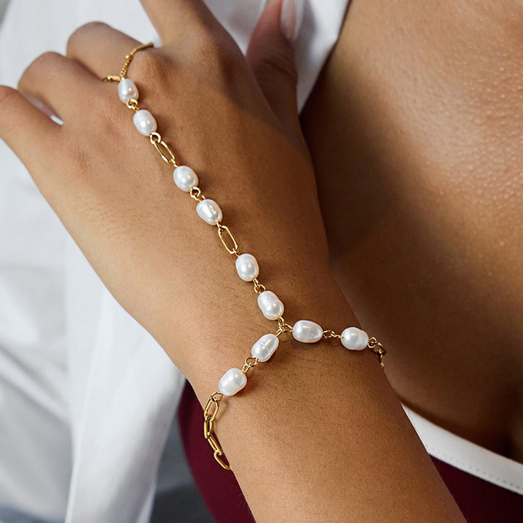 Bracelet chaîne doré relié à un anneau, orné de perles blanches ovales sur main féminine.