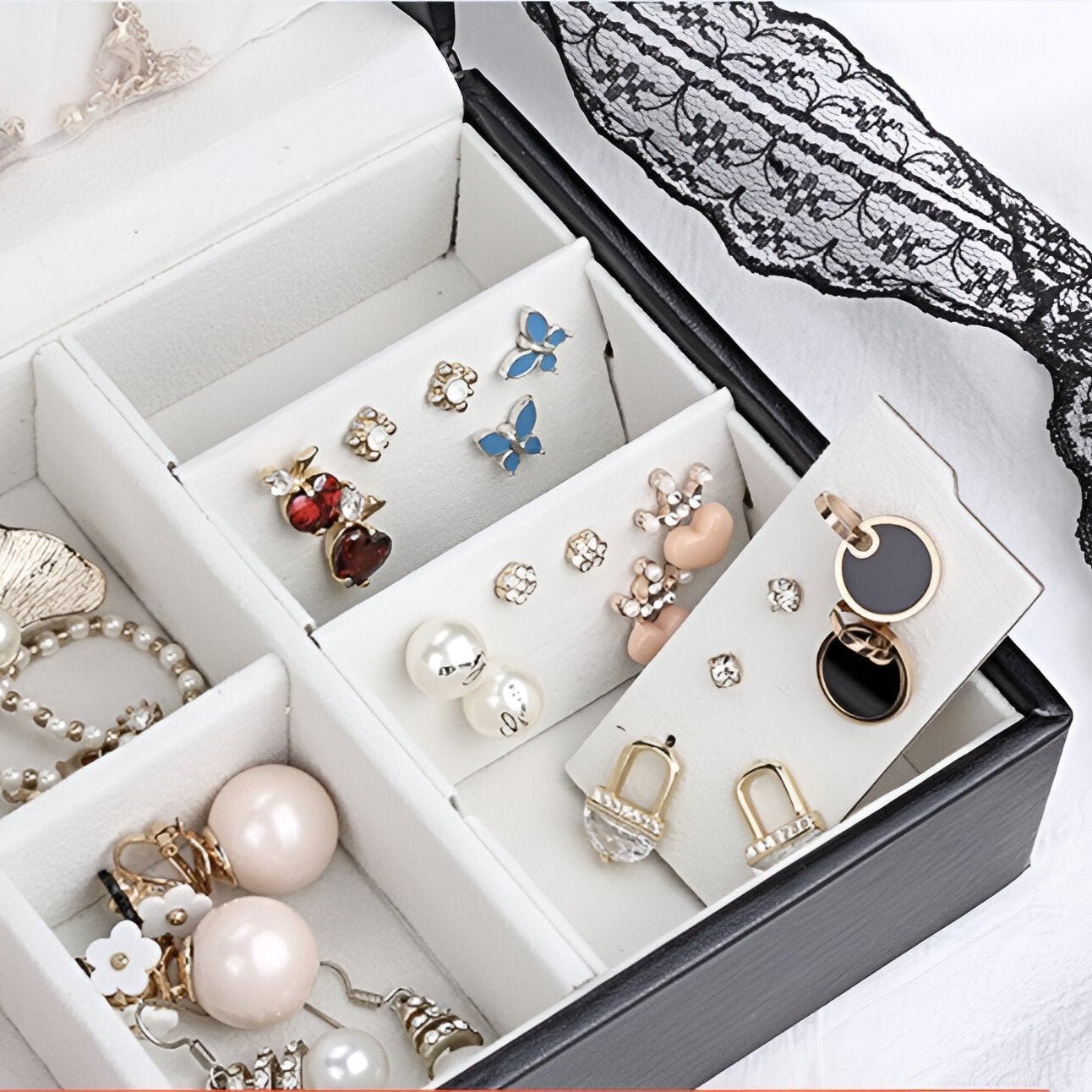 Boîte à bijoux noire ouverte avec compartiments rembourrés et boucles d'oreilles variées, dentelle noire en arrière-plan.