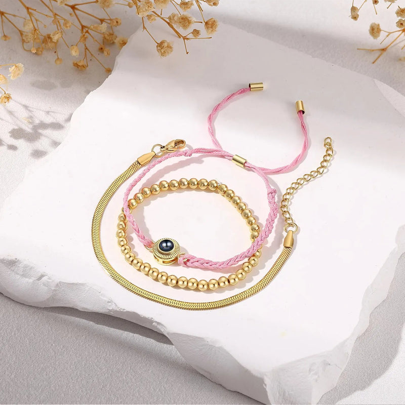 Bracelet trio en acier inoxydable avec perles dorées, cordon tressé rose et pendentif œil bleu.