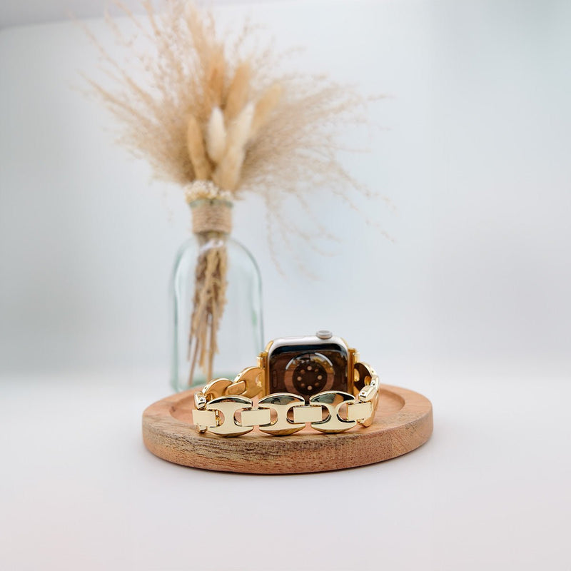 Bracelet Apple Watch femme à mailles larges en métal doré, design moderne, posé sur plateau en bois clair.