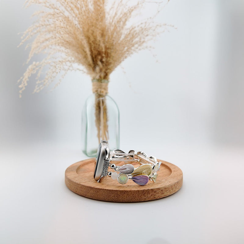Bracelet Apple Watch femme en métal argenté avec breloques en forme de plumes colorées, posé sur un socle en bois rond.