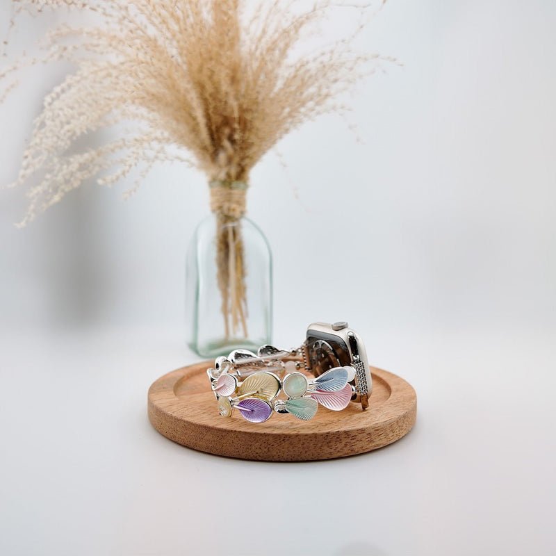 Bracelet Apple Watch pour femme avec breloques plume pastel multicolore sur socle bois rond.