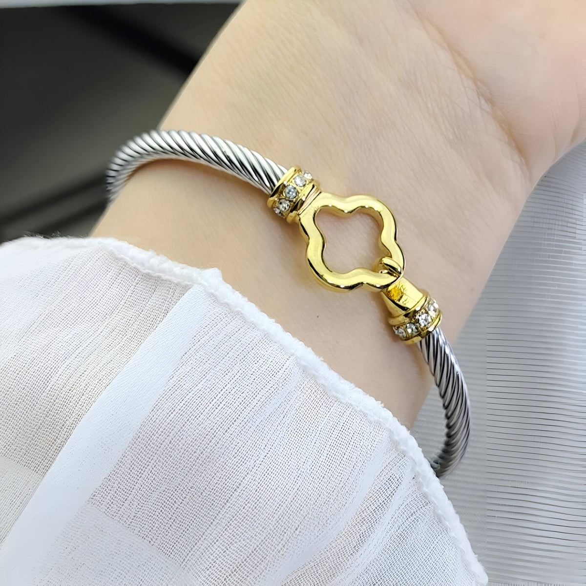 Bracelet câble acier inoxydable argenté avec fermoir doré en forme de trèfle et détails strassés.