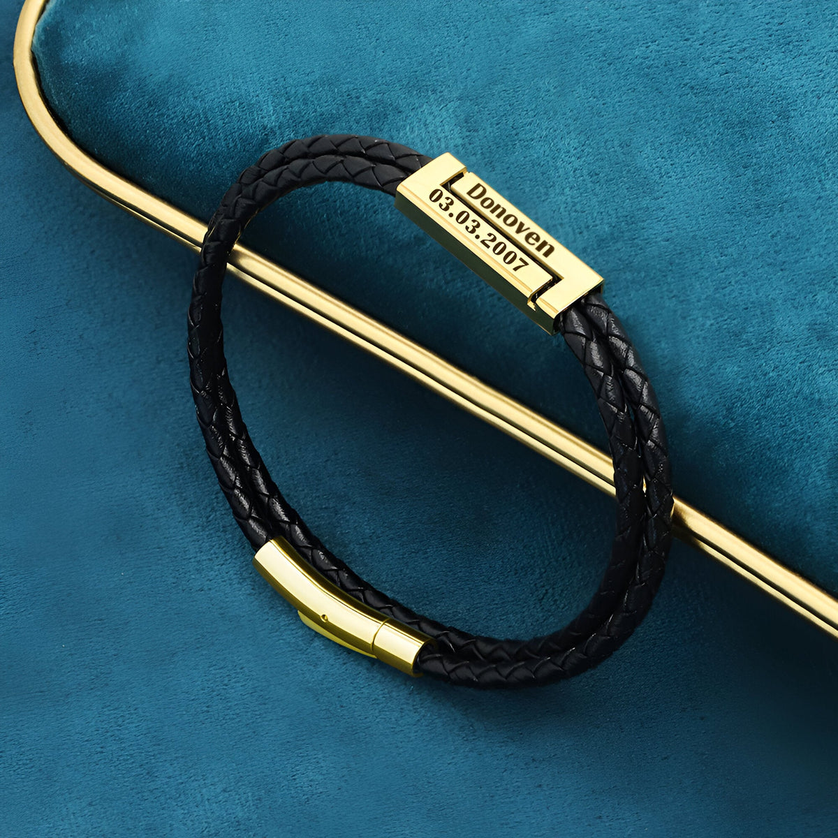 Bracelet cordon noir en cuir tressé avec plaque argentée gravée et fermoir magnétique doré.