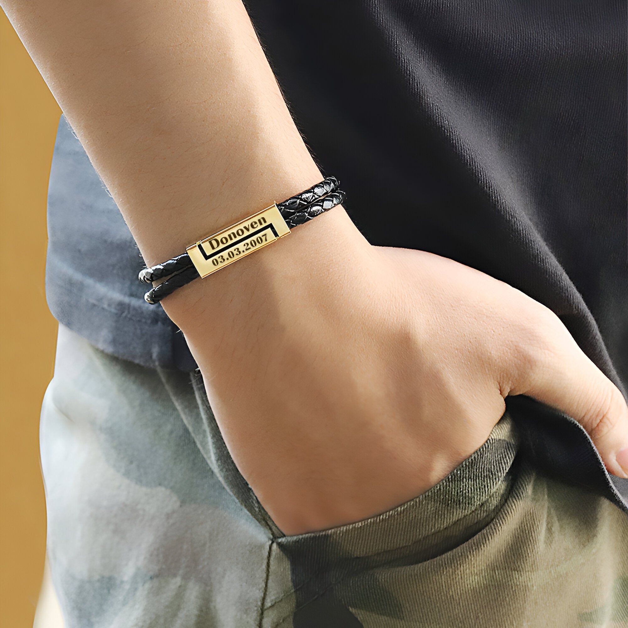 Bracelet cordon noir en cuir avec plaque métallique dorée gravée, porté au poignet.