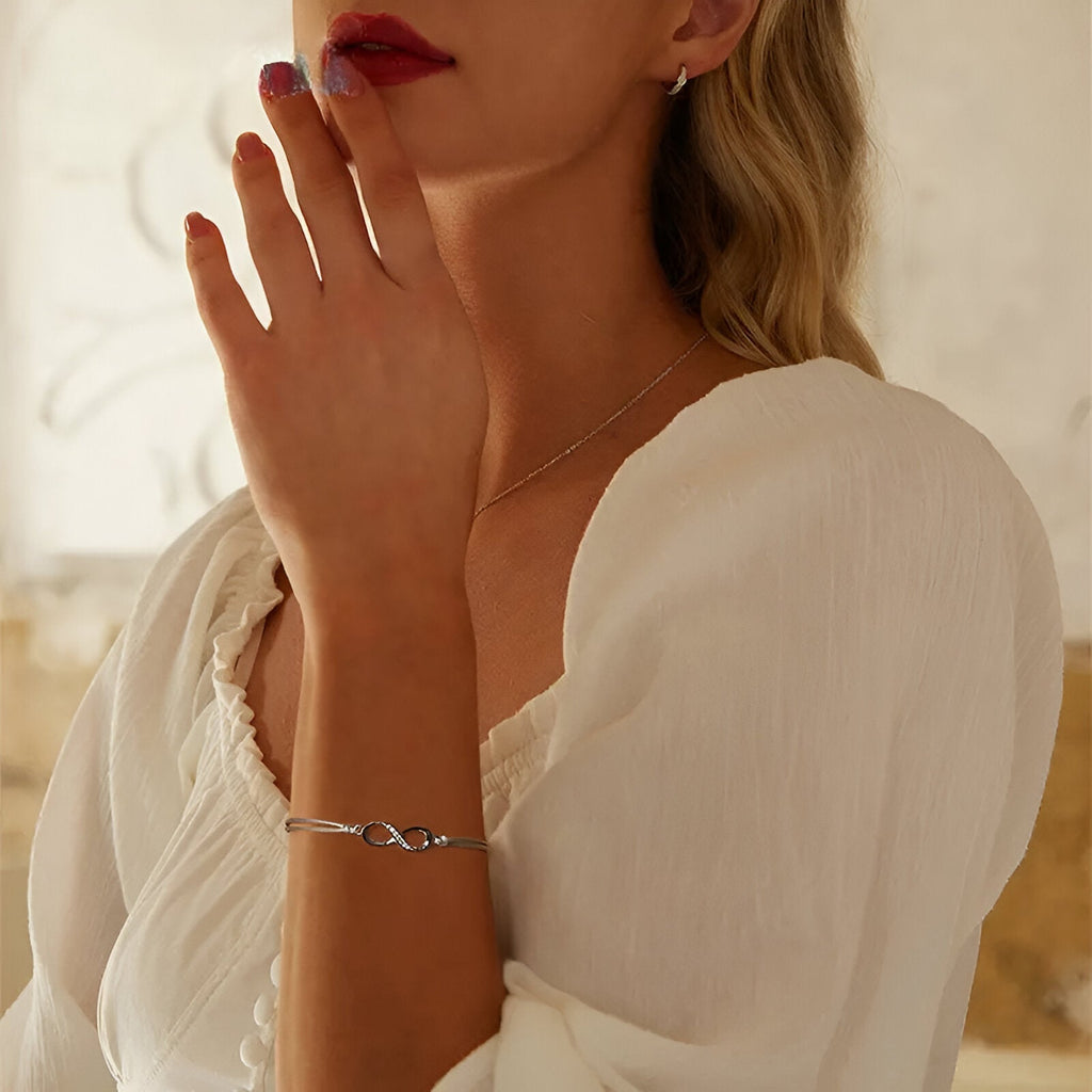 Bracelet cordon argent avec charm infini, porté sur poignet féminin avec blouse blanche fluide.
