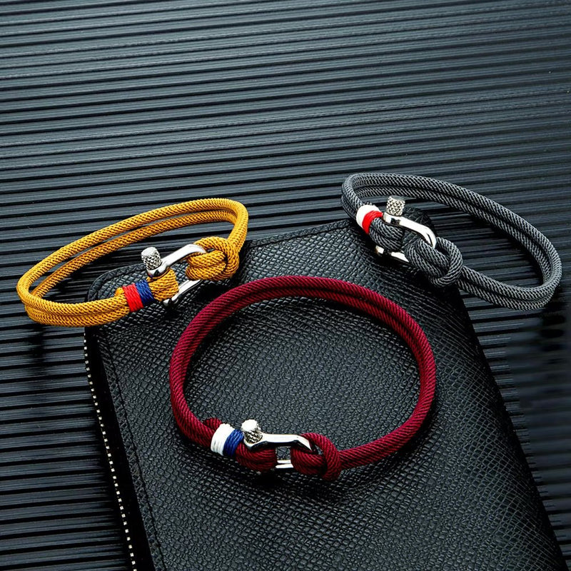 Trois bracelets cordon homme avec manilles acier inoxydable, couleurs jaune, bordeaux et noir, sur surface texturée noire.