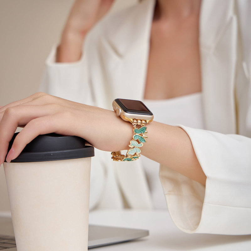 Bracelet Apple Watch bleu à motif papillons en métal doré porté au poignet féminin avec veste blanche.