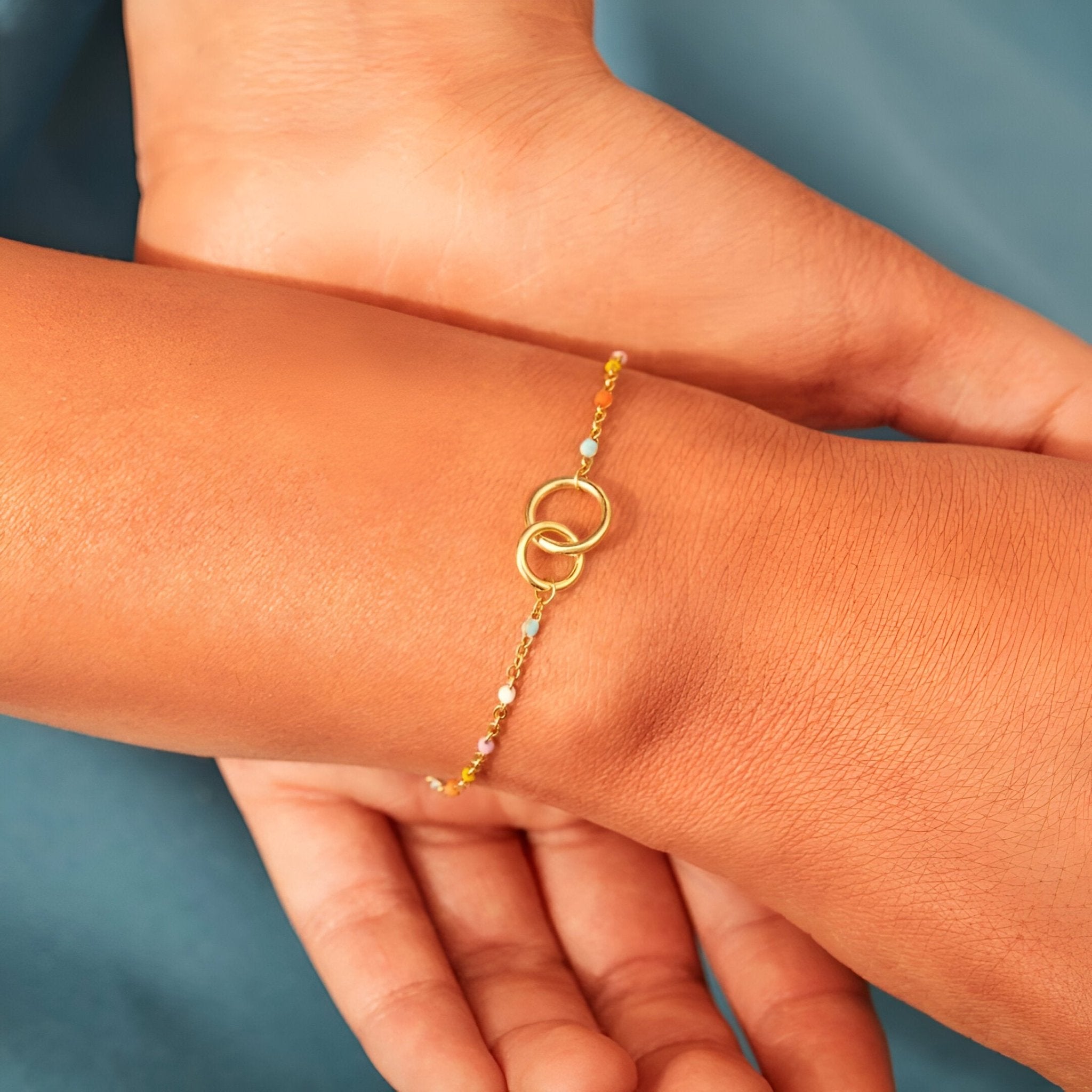Bracelet fin en argent doré avec deux anneaux entrelacés et petites perles colorées.