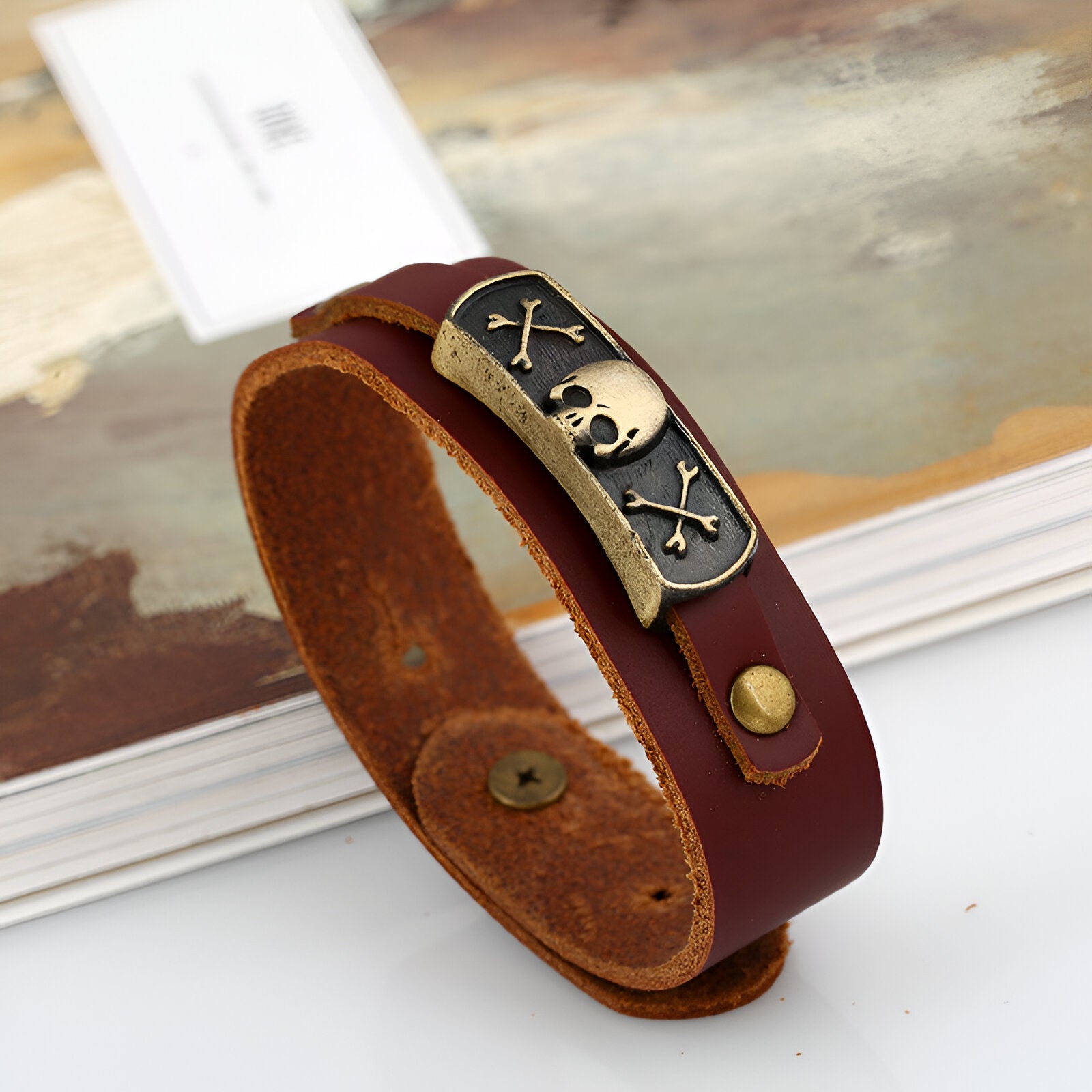Bracelet en cuir marron avec plaque métallique noire ornée d'une tête de mort dorée et d'os croisés.