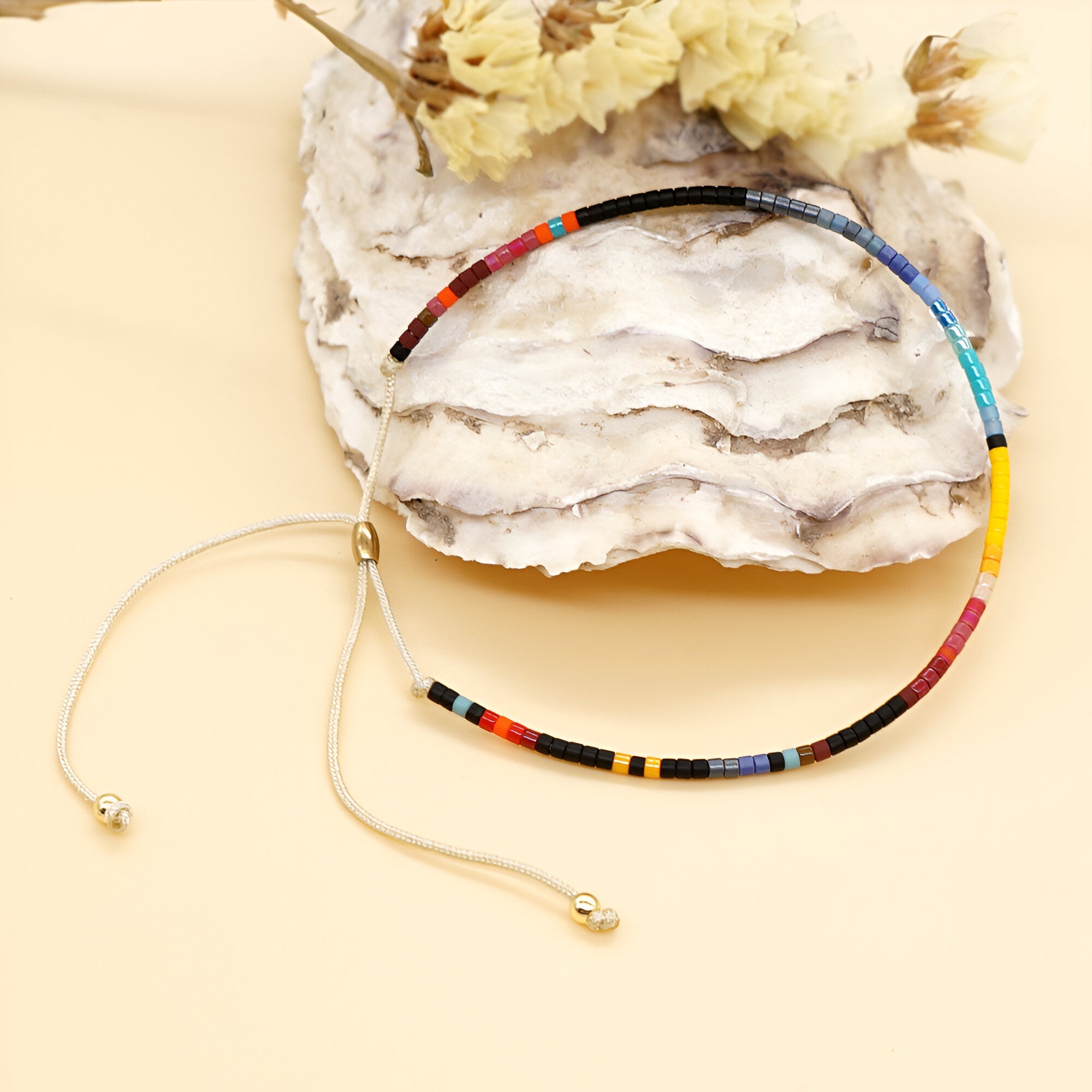 Bracelet ajustable en perles fines multicolores style Kumiko posé sur coquillage avec fleurs sèches en arrière-plan.