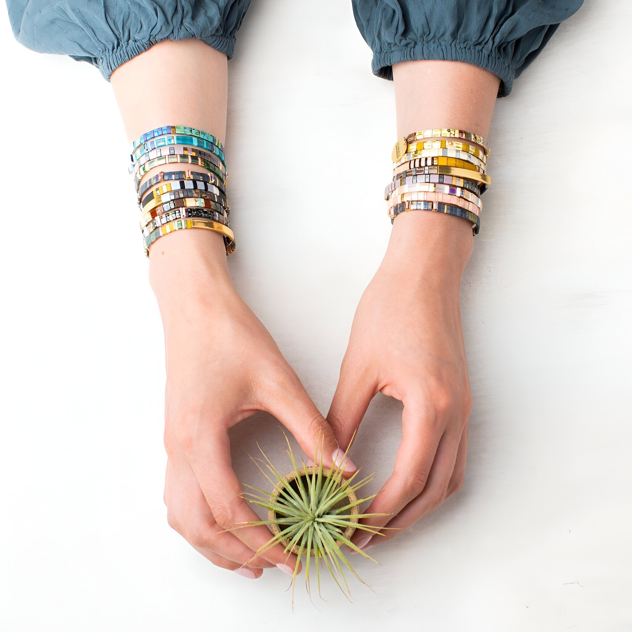 Bracelets en perles japonaises bleu et doré portés sur poignets féminins tenant une plante aérienne.