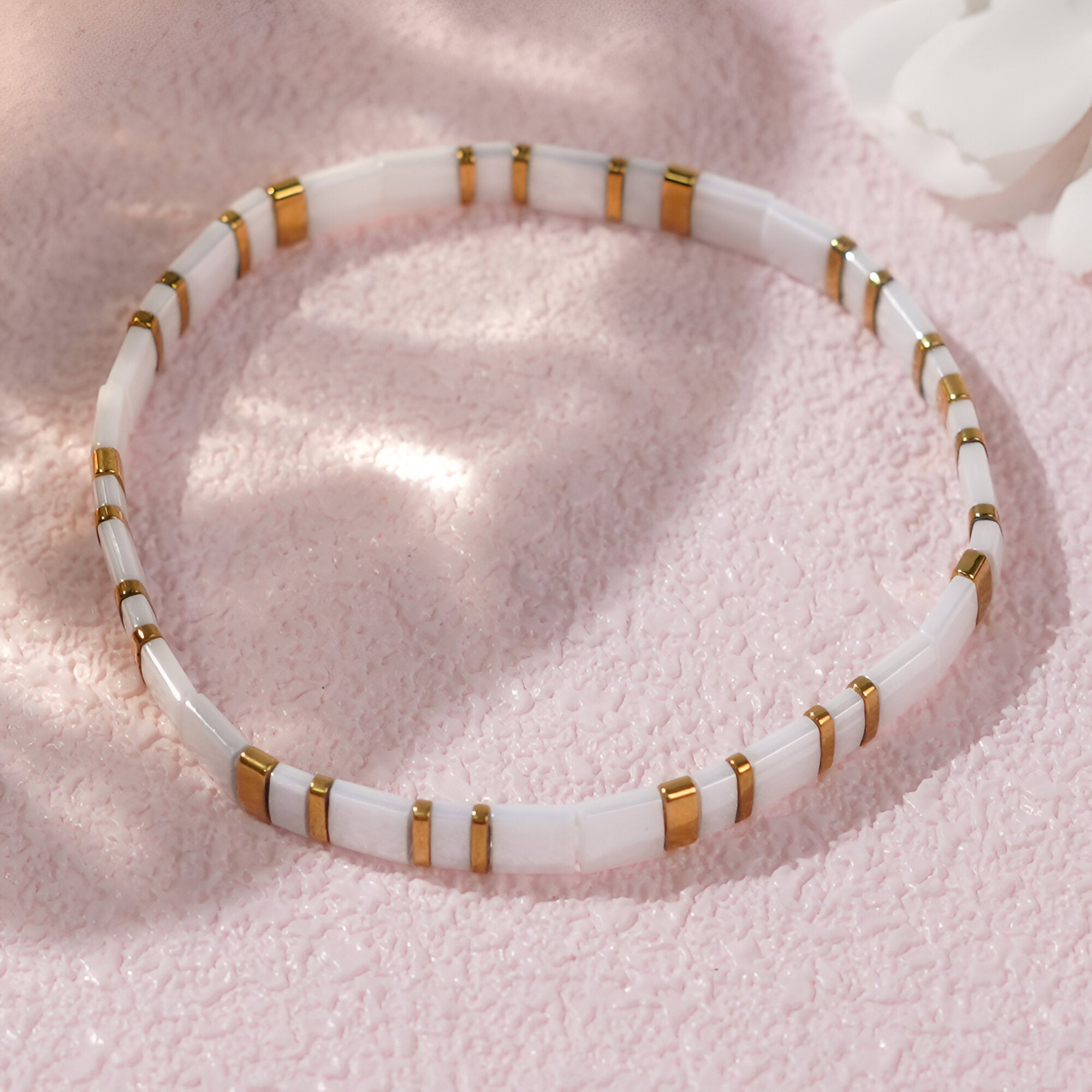 Bracelet féminin en perles blanches japonaises cylindriques avec anneaux dorés sur fond rose doux.