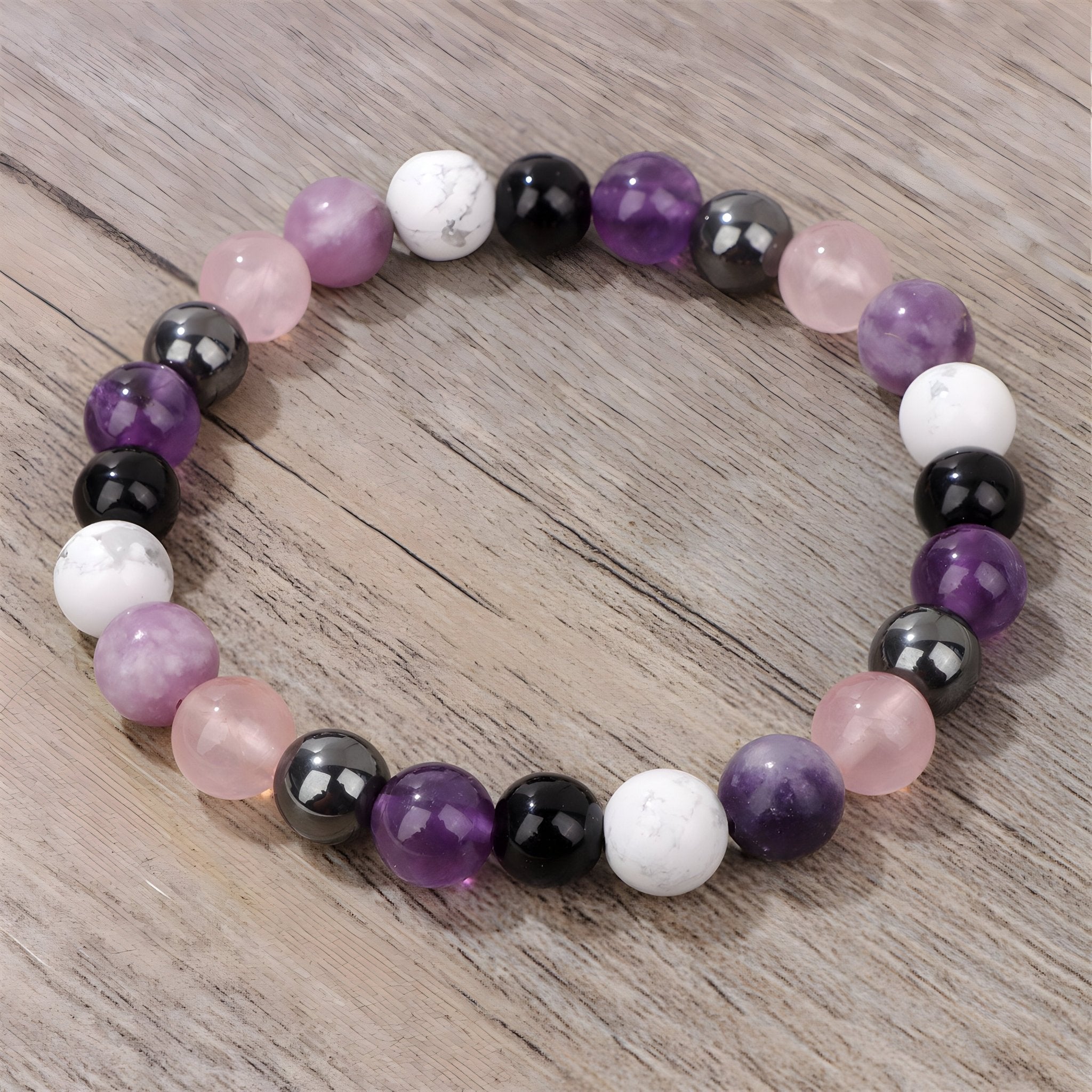 Bracelet élastique à perles rondes en pierres naturelles violettes, blanches, noires et roses sur fond bois clair.