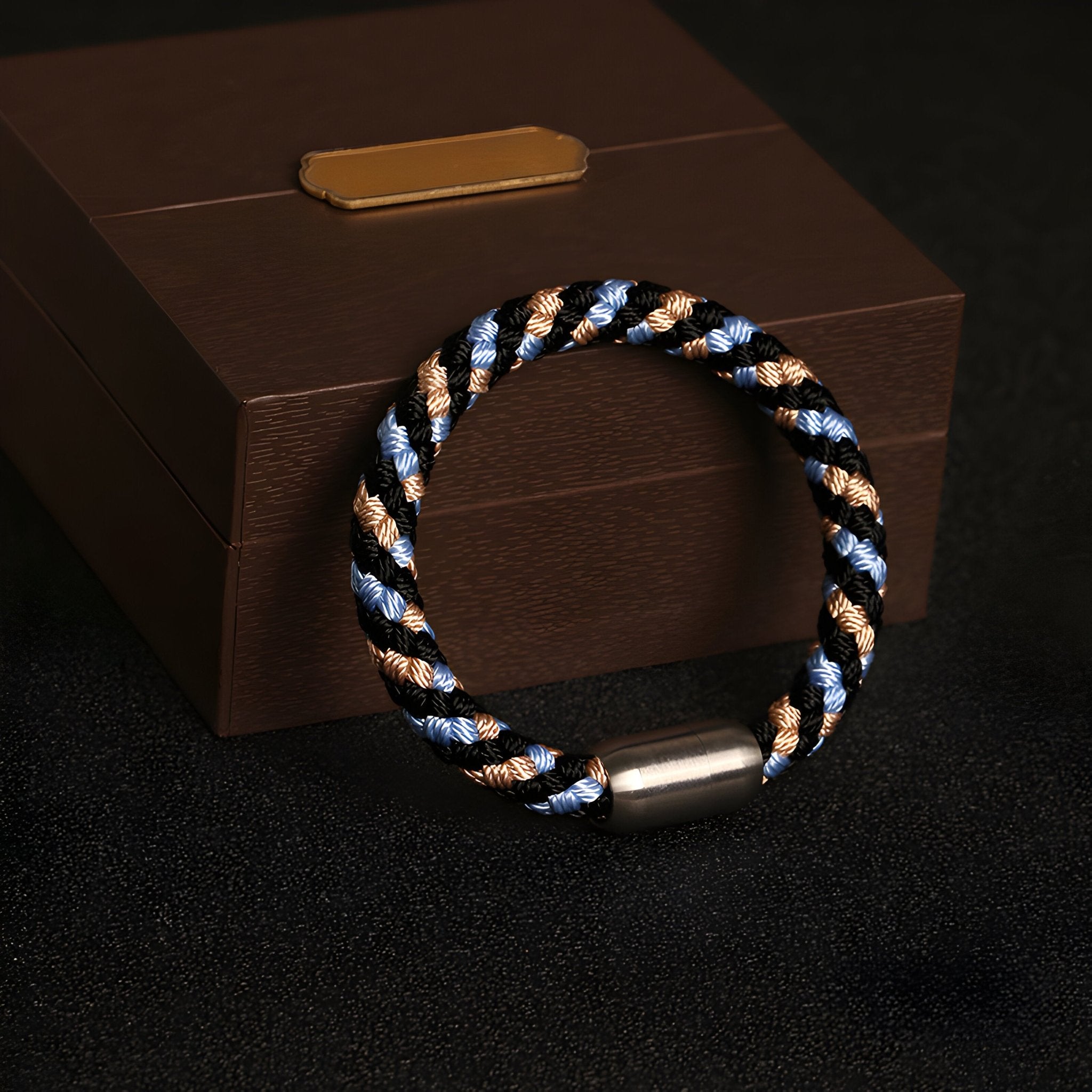 Bracelet tressé polyester noir, bleu et beige avec fermoir métallique argenté posé sur boîte marron.