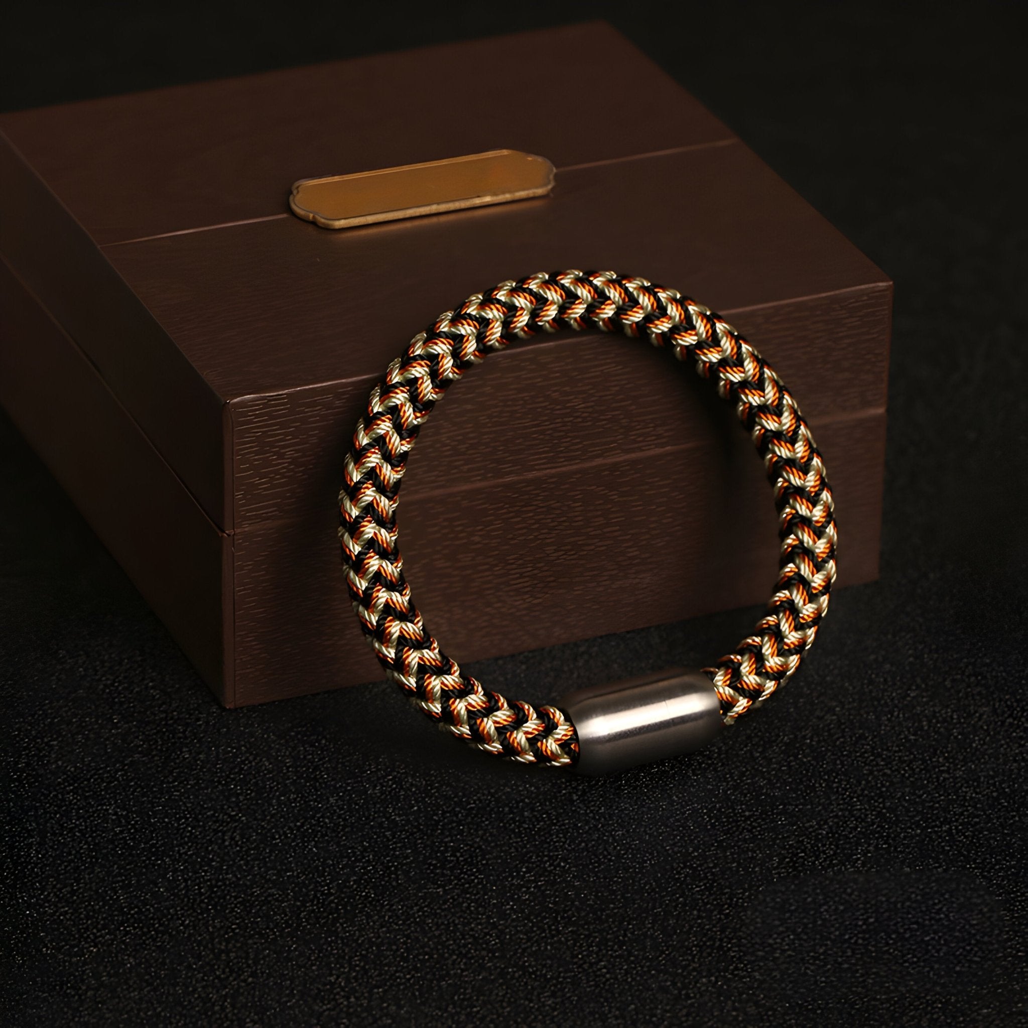 Bracelet tressé en polyester noir, beige et marron avec fermoir argenté sur boîte marron élégante.