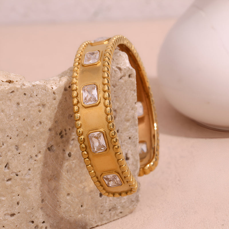 Bracelet jonc doré en acier inoxydable avec pierres carrées et bordure perlée.