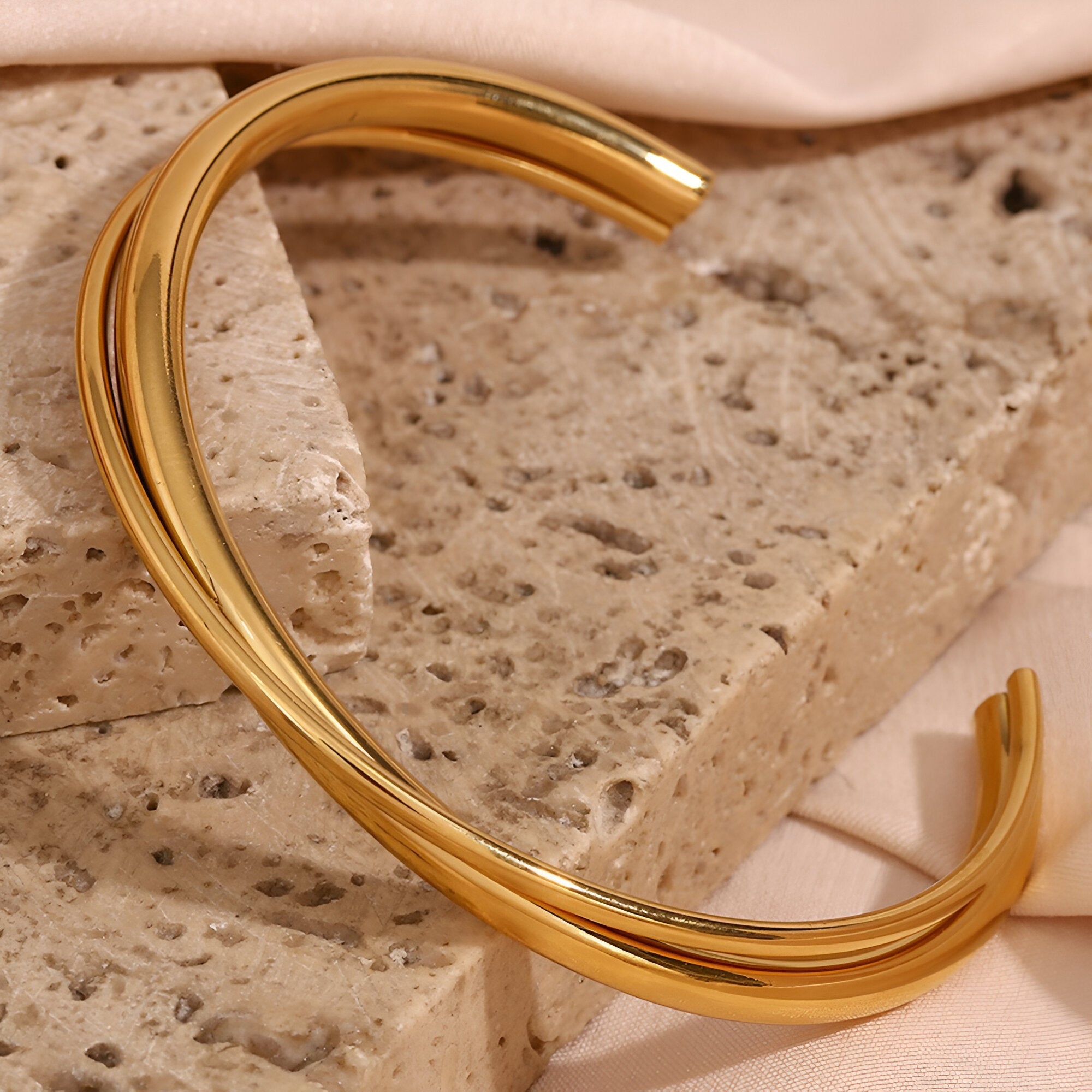 Bracelet jonc duo torsadé en acier inoxydable finition or brillant posé sur pierre beige texturée.