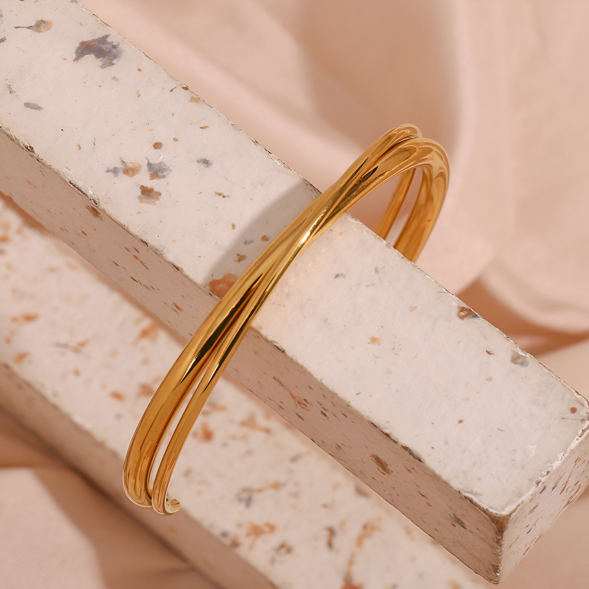 Bracelet jonc duo torsadé en acier inoxydable couleur or brillant posé sur un bloc texturé clair.