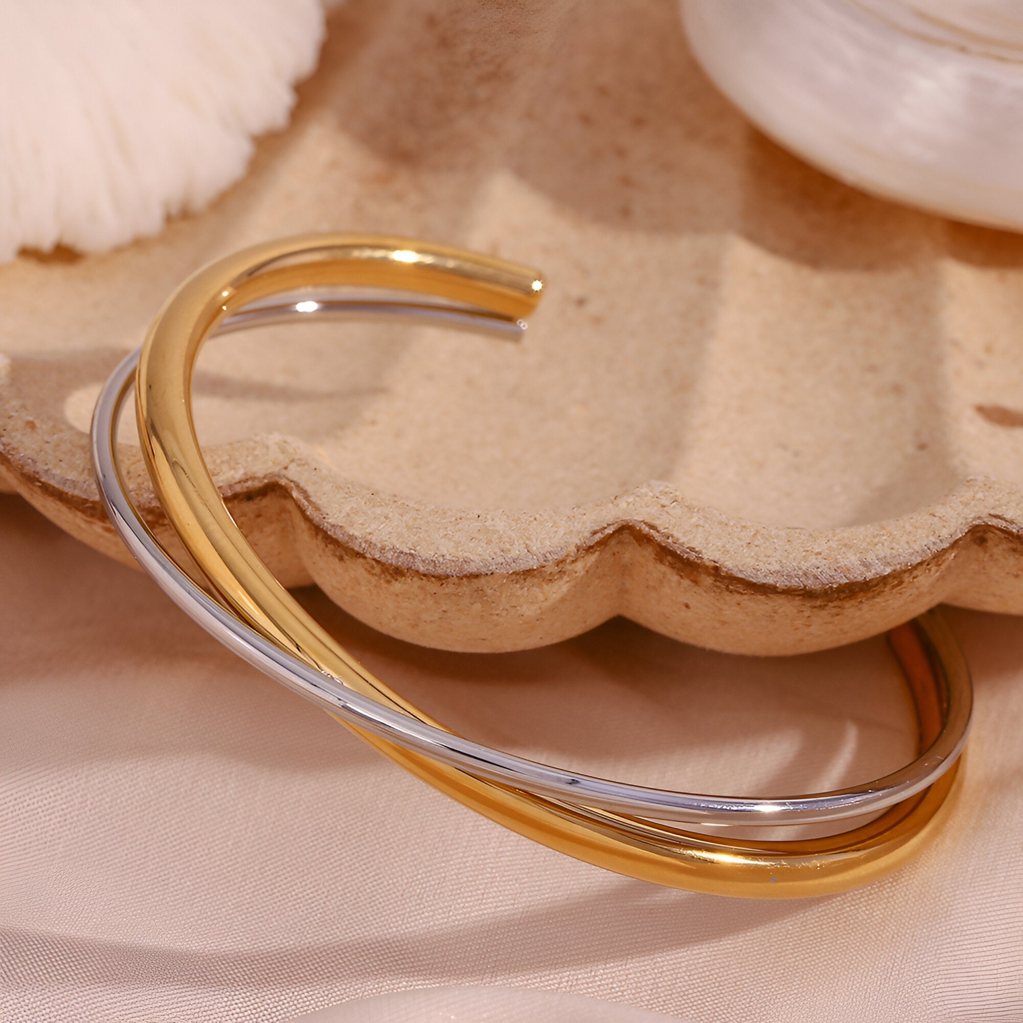 Bracelet jonc duo torsadé en acier inoxydable or et argent modèle Lia posé sur plateau coquillage beige.