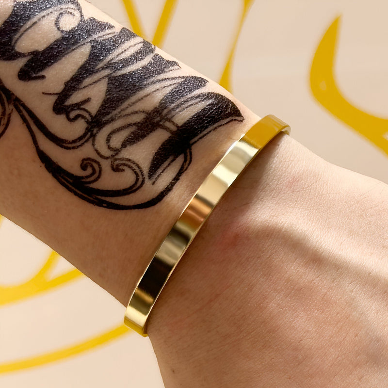 Bracelet jonc fin en acier inoxydable doré, finition brillante, porté sur un poignet tatoué.