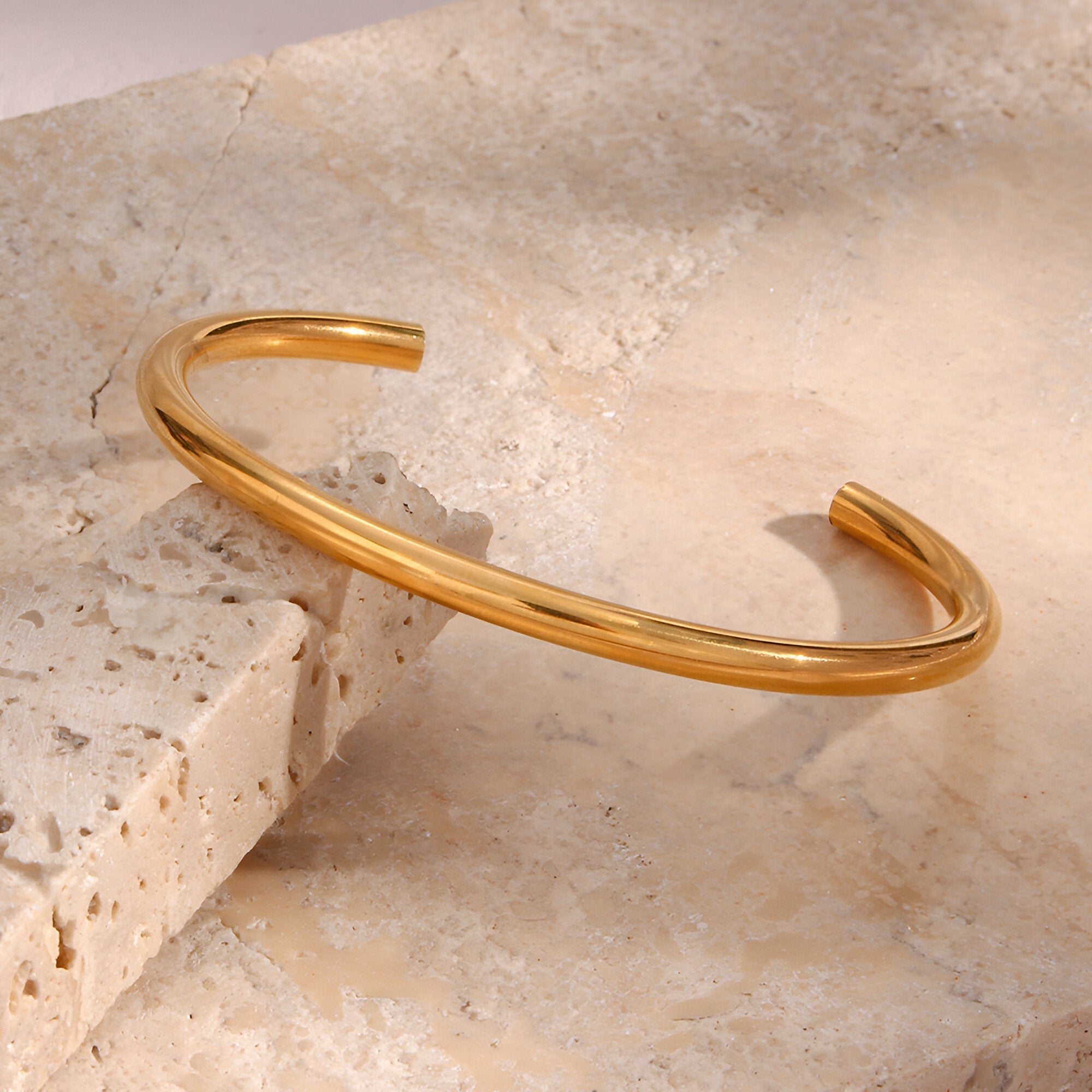 Bracelet jonc fin en acier inoxydable doré au design épuré posé sur pierre beige texturée.