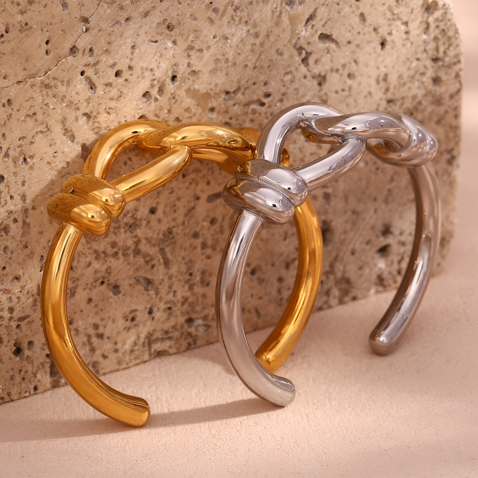 Bracelet jonc en acier inoxydable or, design nœud d'aigui, modèle Cassiopée et bracelet argenté même modèle posé sur fond.