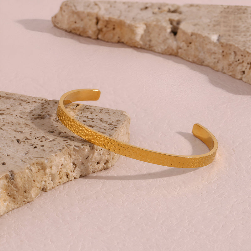 Bracelet jonc doré en acier inoxydable avec finition martelée posée sur pierres beiges texturées.