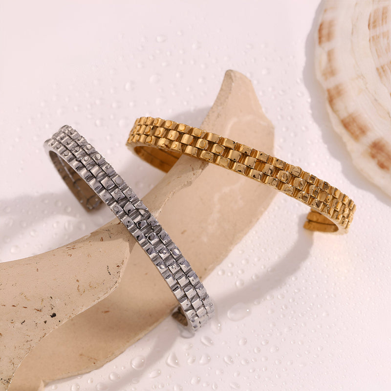 Bracelet jonc maille gourmette en acier inoxydable or et argent posé sur décor naturel sable et coquillage.