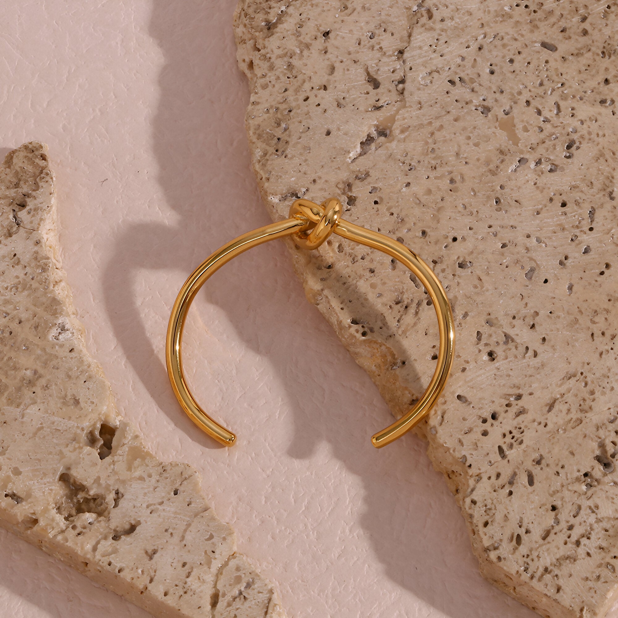 Bracelet jonc or en acier inoxydable avec nœud infini élégant sur fond pierre beige.