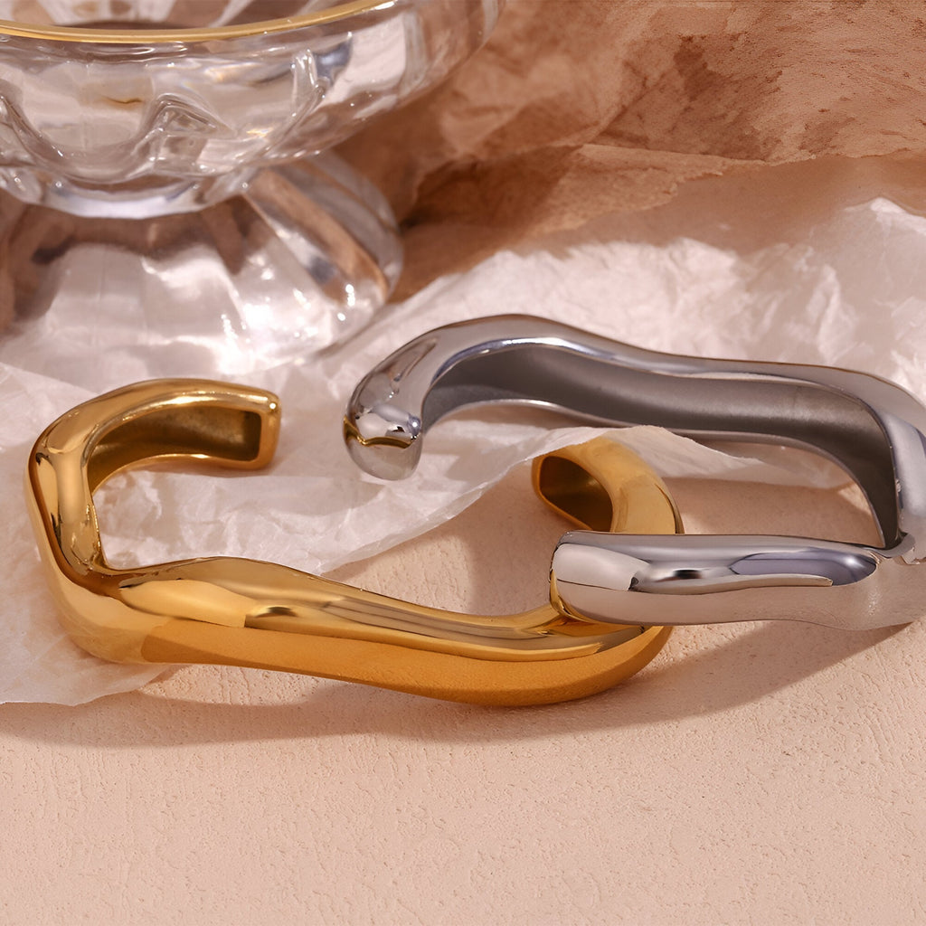 Deux bracelets joncs ondulés en acier inoxydable, un doré et un argenté, sur fond beige.