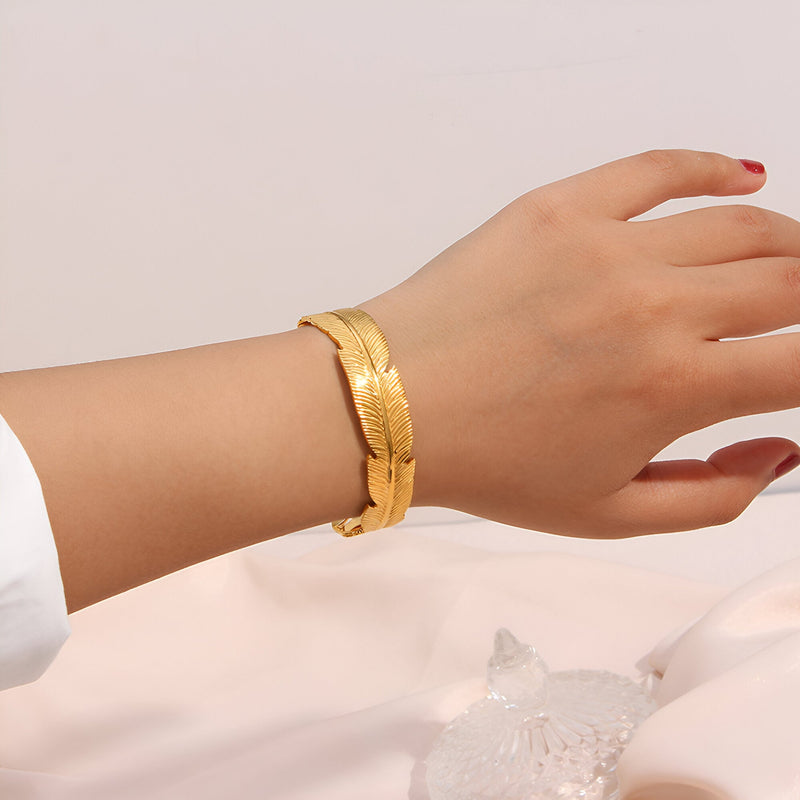 Bracelet jonc doré en acier inoxydable orné d'une plume sculptée, porté au poignet féminin.