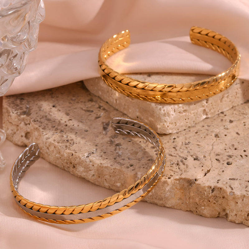 Deux bracelets joncs tressés en acier inoxydable, finition or et argent, posés sur pierre beige.