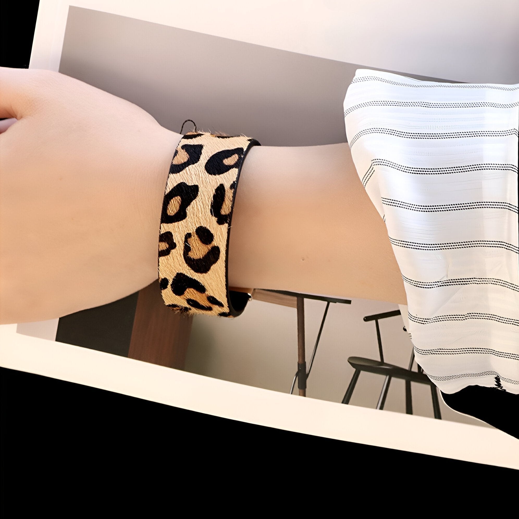 Bracelet manchette beige à motifs léopard noirs porté sur poignet avec chemise à rayures blanches et grises.