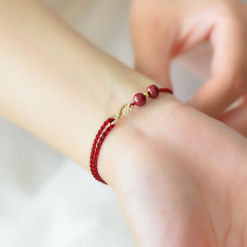 Bracelet minimaliste rouge bordeaux en fil ciré avec pierres de cinabre et motif infini doré sur poignet féminin.