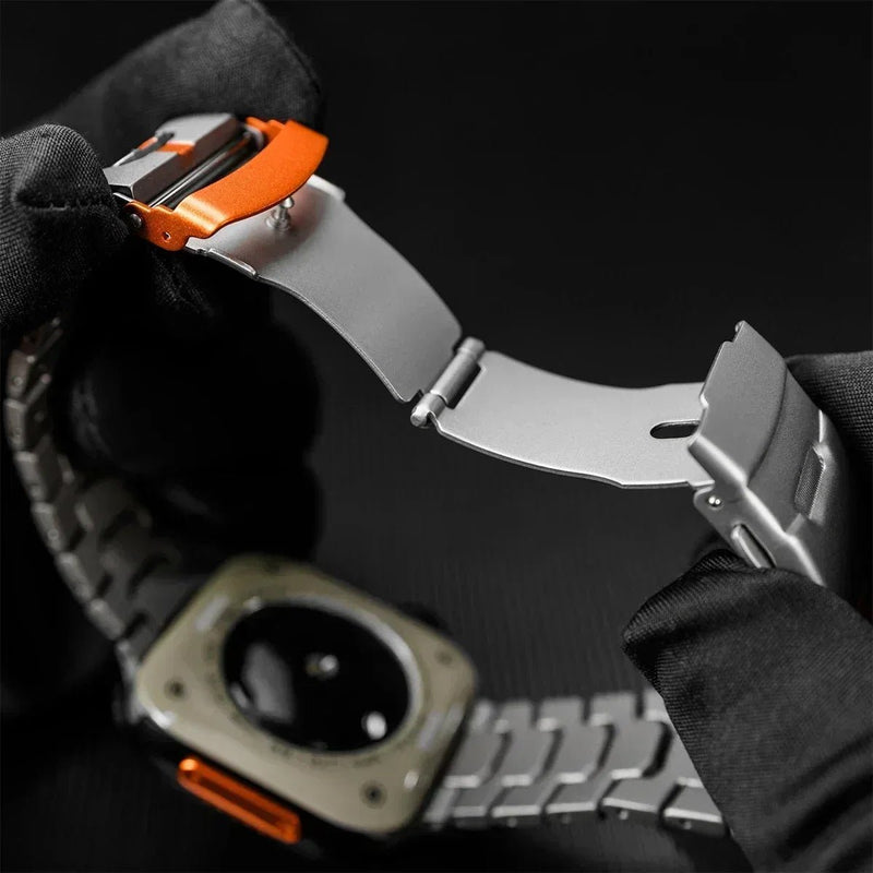 Bracelet montre Apple en titane gris avec maille moderne, boucle déployante argentée et ligne orange vive distinctive.