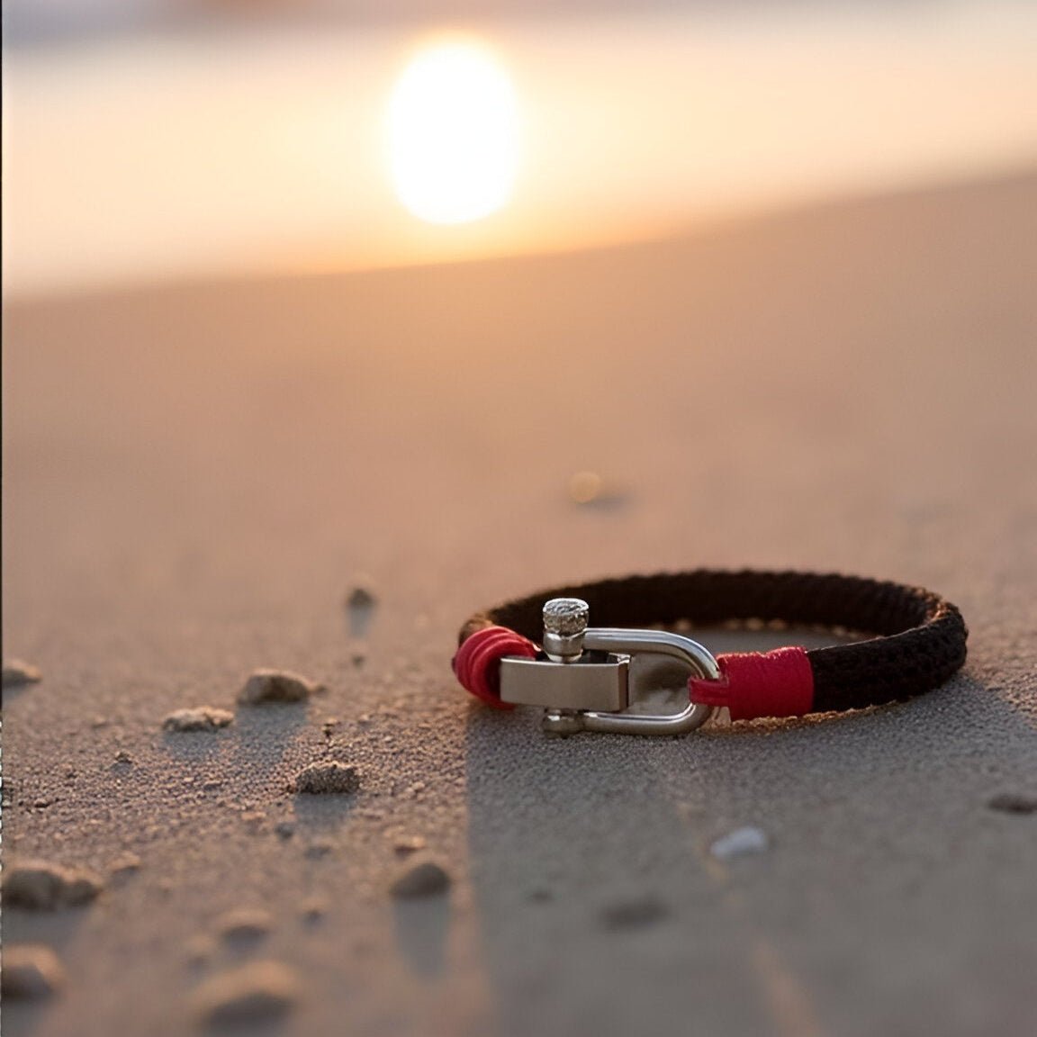 Bracelet homme en corde noire et rouge avec fermoir mousqueton argenté posé sur sable au coucher du soleil.