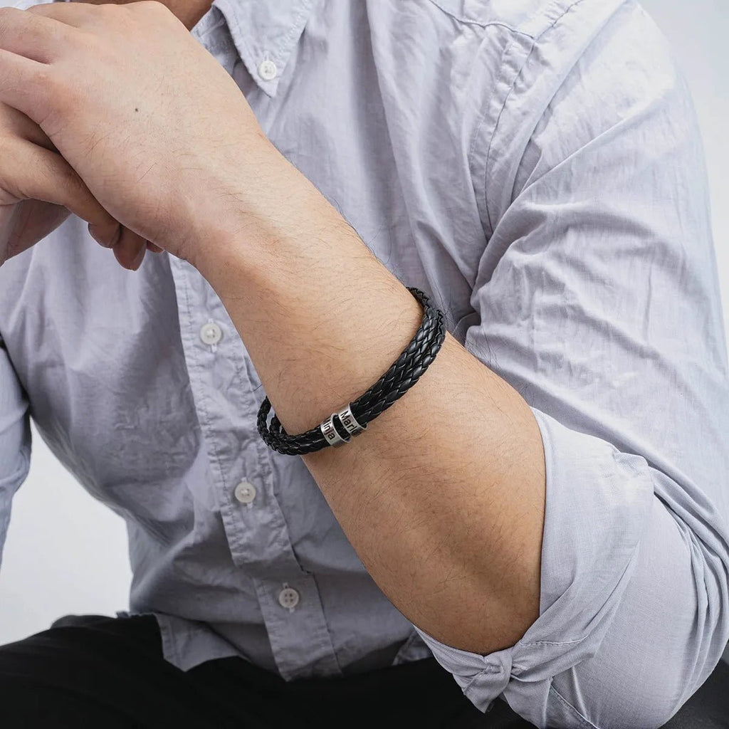 Bracelet en cuir noir triple tour tressé avec deux anneaux argentés gravables porté sur poignet homme.