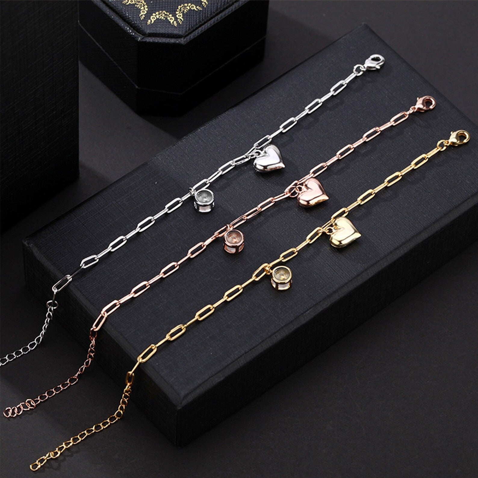 Trois bracelets personnalisés en titane avec charms cœur en or, argent et or rosé sur fond noir.