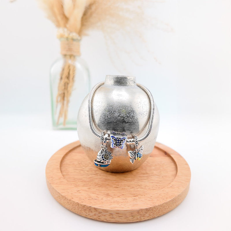 Bracelet argenté avec charms papillon bleu et breloques scintillantes sur présentoir métallique rond.