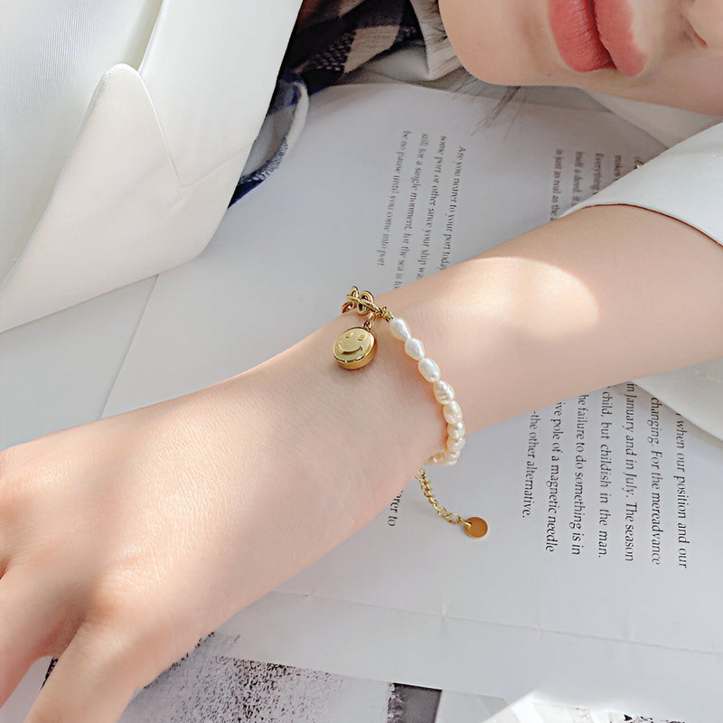 Bracelet femme en acier doré avec perles blanches et charm smiley en métal sur poignet.