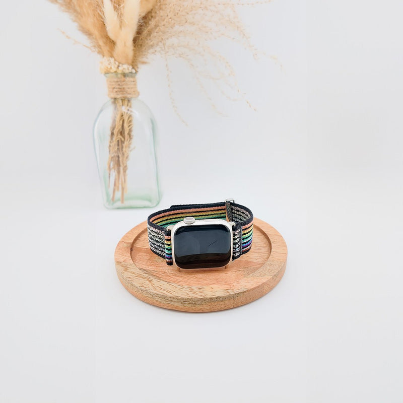 Bracelet Apple Watch en nylon noir avec rayures colorées et fermeture velcro, posé sur un plateau en bois rond.