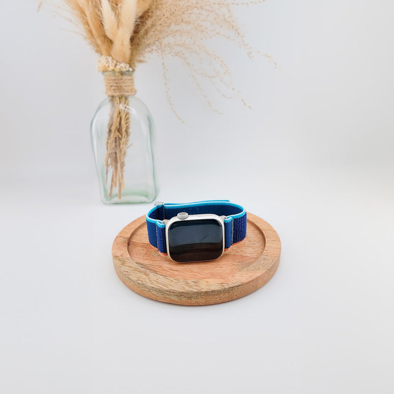 Bracelet Apple Watch en nylon bleu résistant avec fermeture velcro, modèle Elios, posé sur un support bois rond.