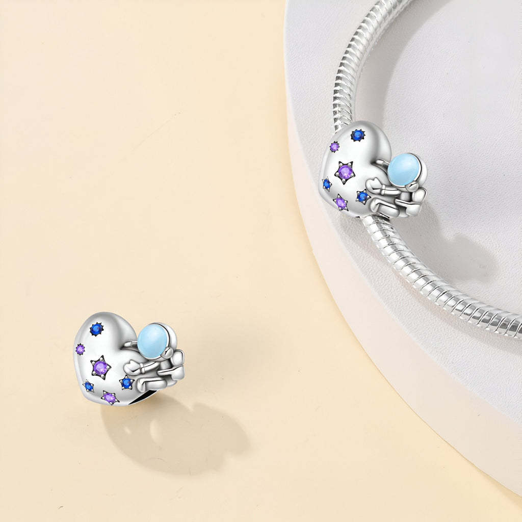 Charm argenté en forme de cœur avec astronaute bleu et étoiles violettes, style fantaisie.