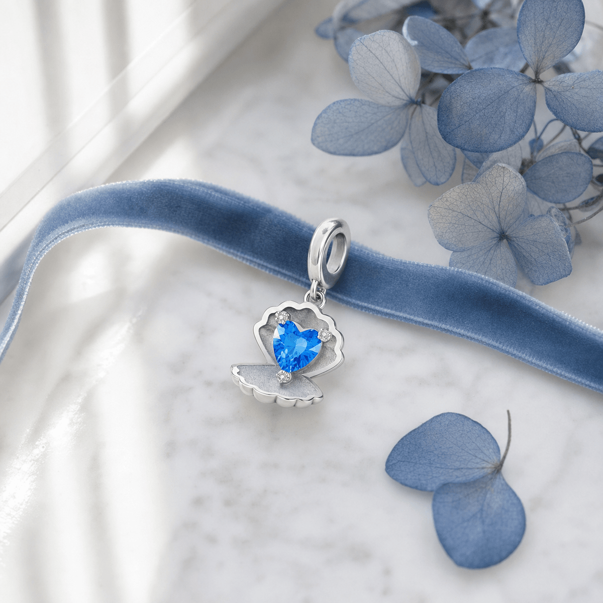 Charm Conchilia coquillage cœur bleu posé sur marbre blanc avec ruban velours bleu et pétales d'hortensia
