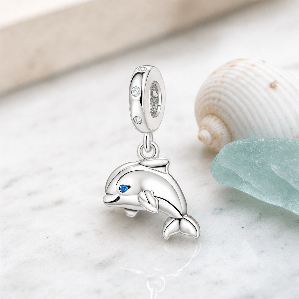 Charm pendentif dauphin argent posé sur marbre blanc avec coquillage et verre de mer