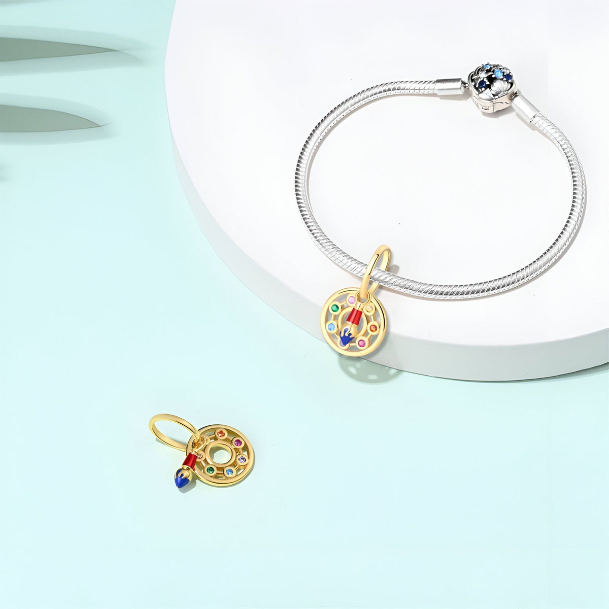 Charm pendentif pinceau doré avec détails colorés et strass bleus sur bracelet argenté fin.