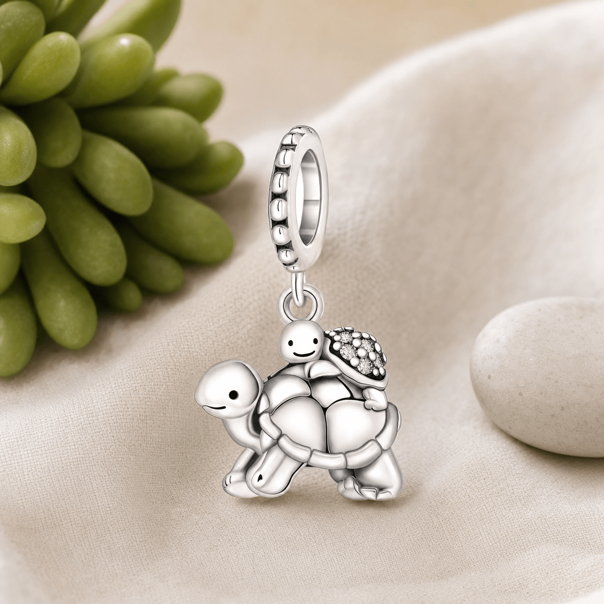 Charm pendentif tortues mère et enfant en argent posé sur tissu lin beige avec plante succulente et galet
