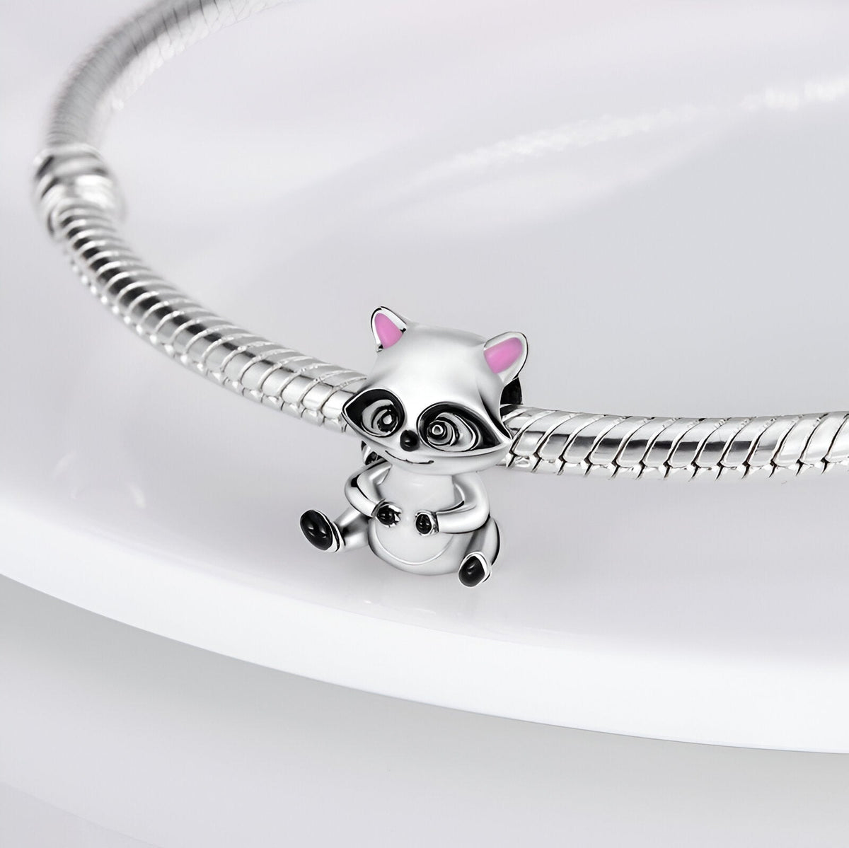 Charm raton laveur argenté avec détails noirs et oreilles roses, suspendu à un bracelet serpent argenté.