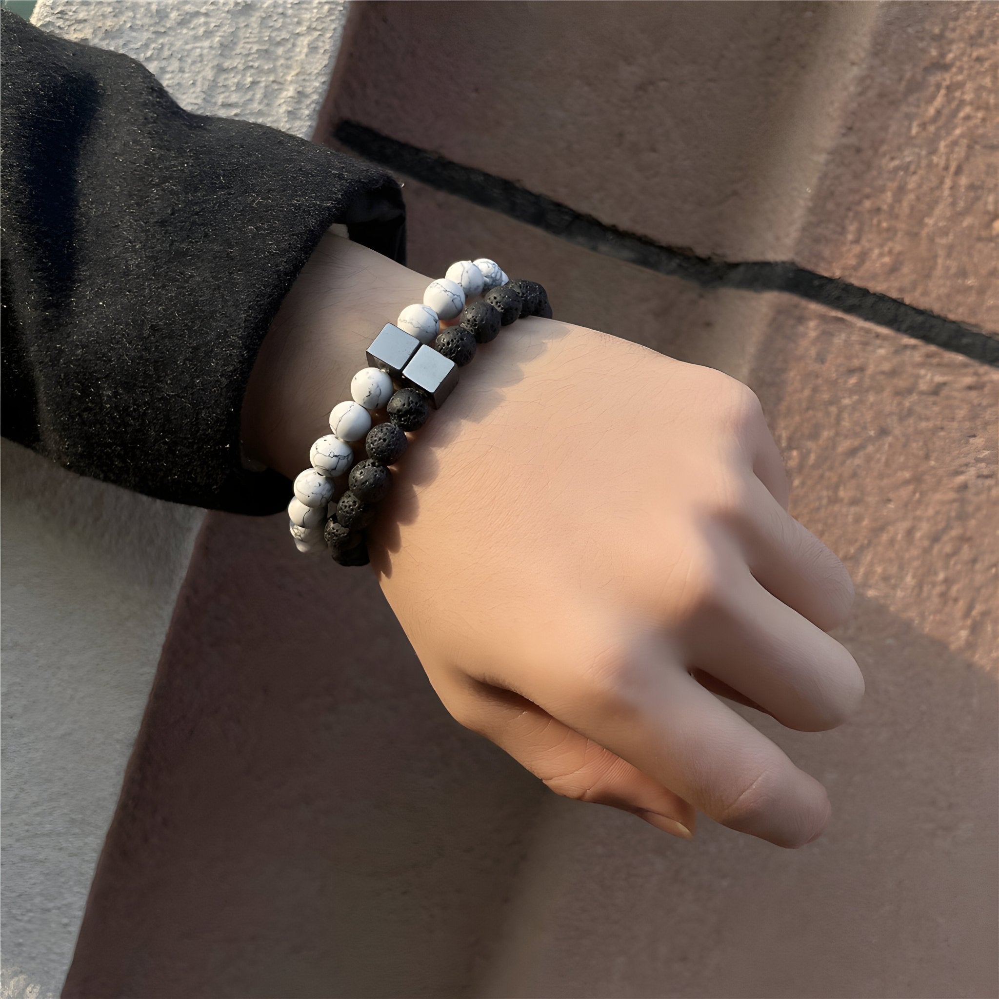 Paire de bracelets élastiques en pierres volcaniques noires et blanches avec perles mates et cube métallique sur poignet.